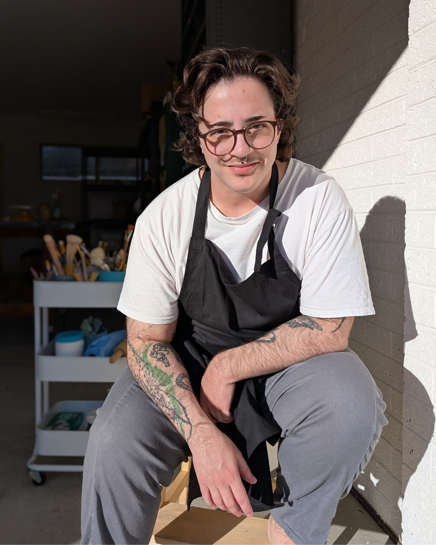 Person wearing a black apron and glasses, sitting against a white wall.